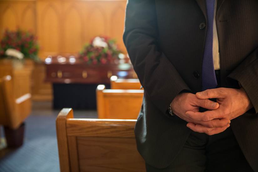 A solemn scene shows a person in a dark suit at a funeral. Wooden pews and a casket adorned with flowers are visible in the background.
